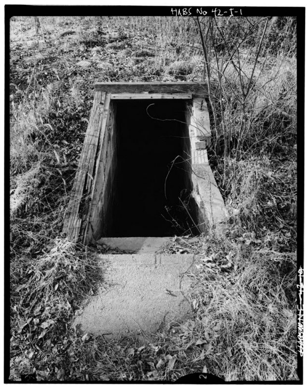 A wood framed rectangular opening in the ground of a high-grass field with concrete stairs leading into a stone walled black hole.