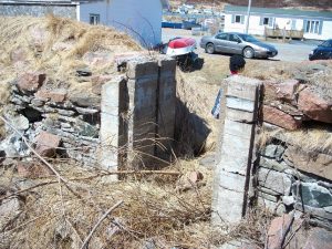 Inside the crumbled stone remains of a root cellar looking out of its partially fallen concrete door frame into the grassy field.