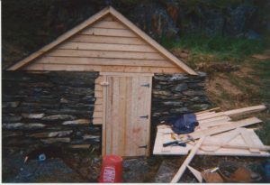 Stone stacked hillside root cellar with an unpainted door and top shed in a grassy hillside.