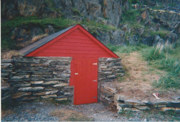 Stone stacked foundations of a hillside root cellar with a red door and top shed in a grassy hillside.