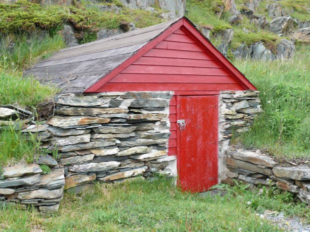Stone stacked hillside root cellar with a red wooden door and red top shed in a grassy field.