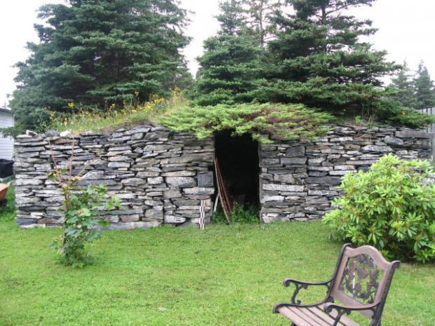 Exterior of a stacked stone root cellar with grass and shrub-covered earth roof.
