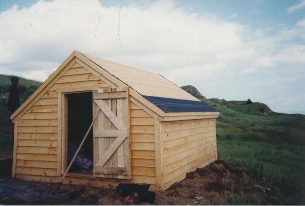 Exterior of the unpainted root cellar top shed in a grassy field.