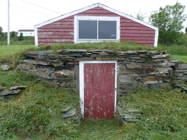Stone stacked hillside root cellar with a red wooden door and stop shed on a grassy hillside.