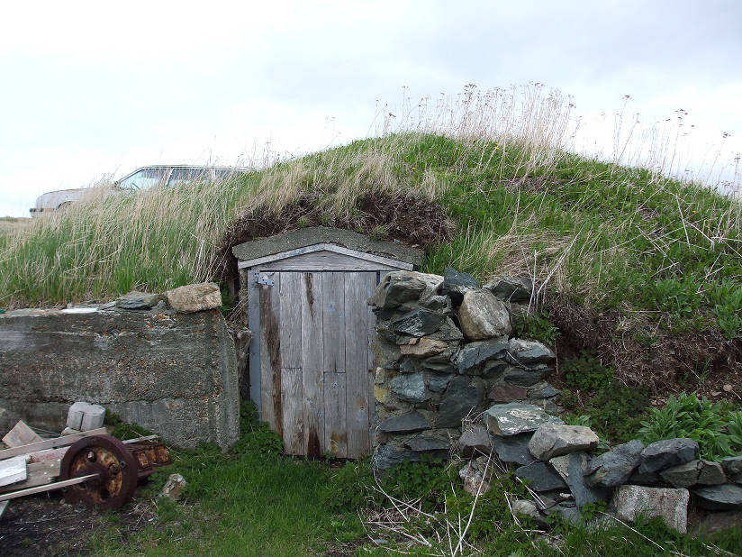 Exterior of a hillside root cellar with concrete wall (left), a wooden entrance door (center) and a pile of stacked stones (right).