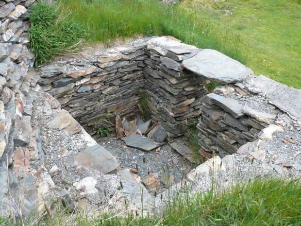 Stacked flat stone wall foundation built into a grass-covered hillside.