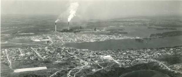 Black-and-white photograph of two cities build around a lake, separated by a vacant lot. In the background, you can see the smelter with three headframes and two smoking chimneys.
