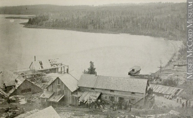 Black-and-white photograph of about fifteen people in front of the four Wright Mine’s wood buildings located along the shores of Lake Timiskaming.