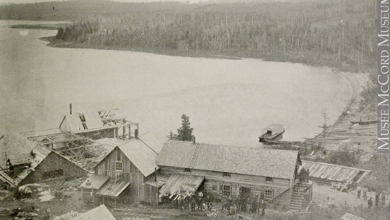 Photographie en noir et blanc d’une quinzaine de personnes devant les quatre bâtiments en planche de la mine Wright situés sur le bord du lac Témiscamingue.