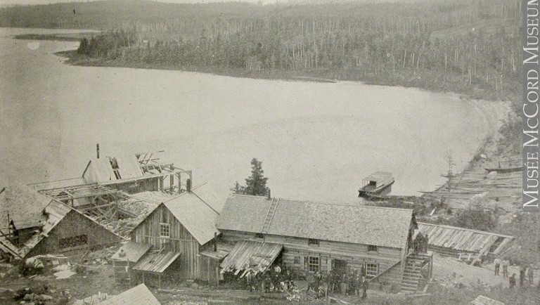 Black-and-white photograph of about fifteen people in front of the four Wright Mine’s wood buildings located along the shores of Lake Timiskaming.