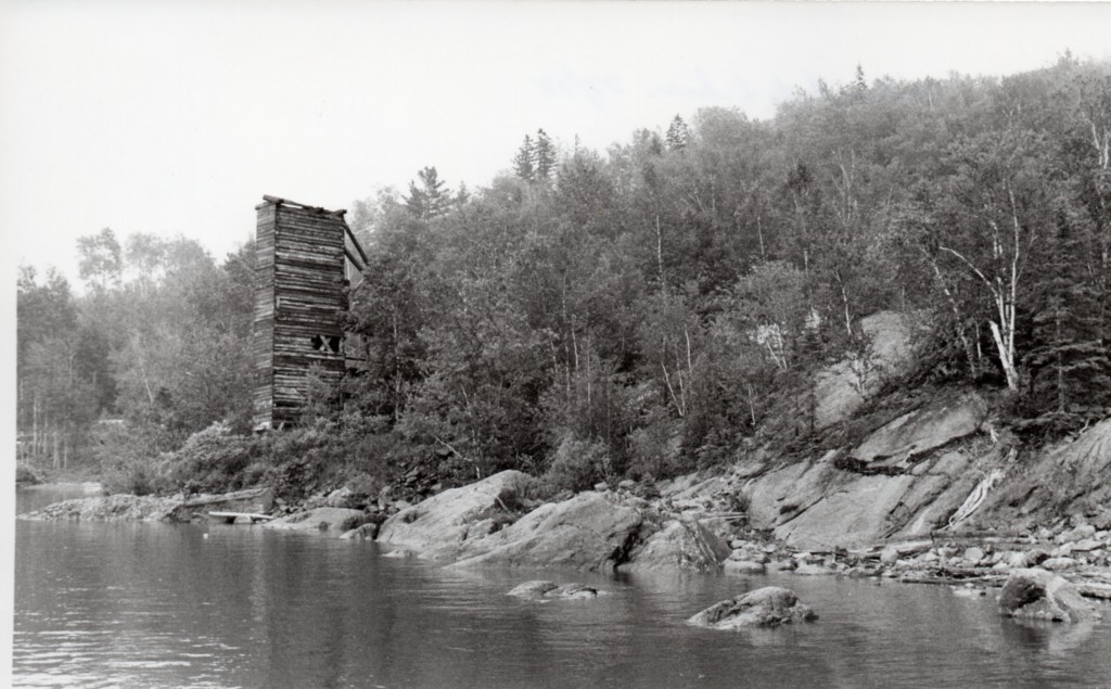 Black-and-white photograph of the shores of Lake Timiskaming, lined with rocks and trees, with a squared high-tower made of wooden planks.