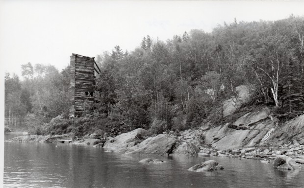 Black-and-white photograph of the shores of Lake Timiskaming, lined with rocks and trees, with a squared high-tower made of wooden planks.