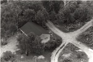 Black-and-white aerial view photograph of a wood building surrounded by trees at the intersection of two gravel paths.