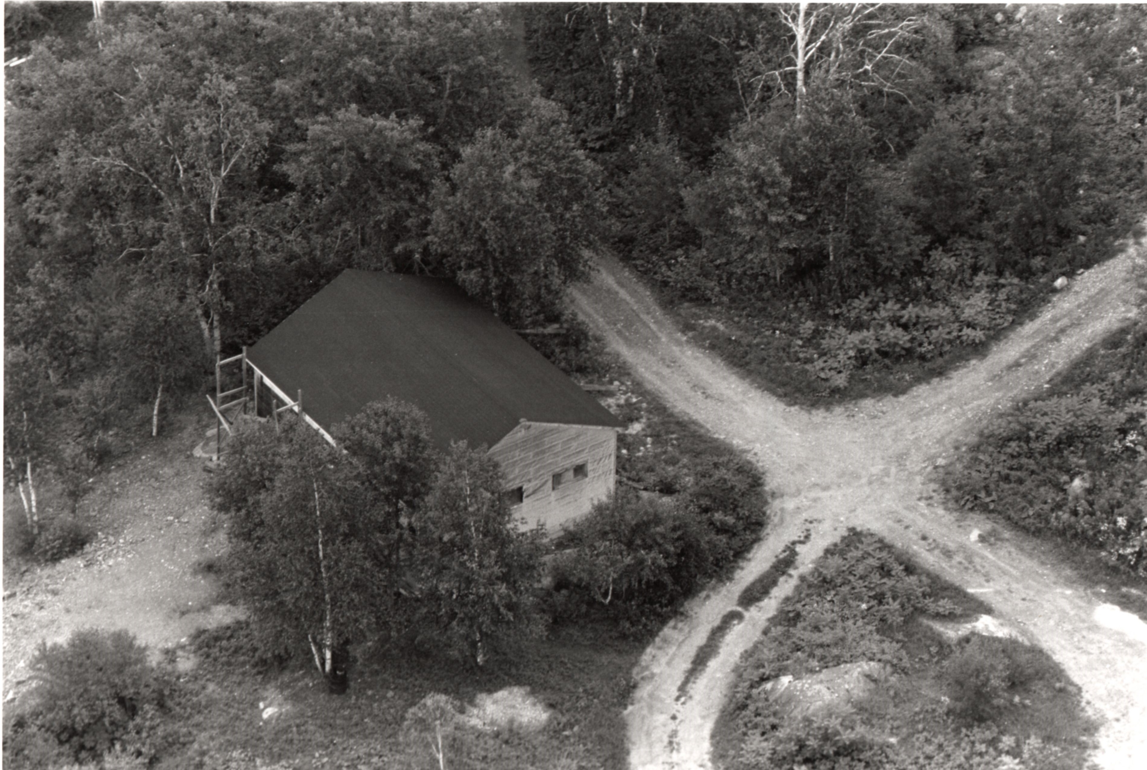 Black-and-white aerial view photograph of a wood building surrounded by trees at the intersection of two gravel paths.