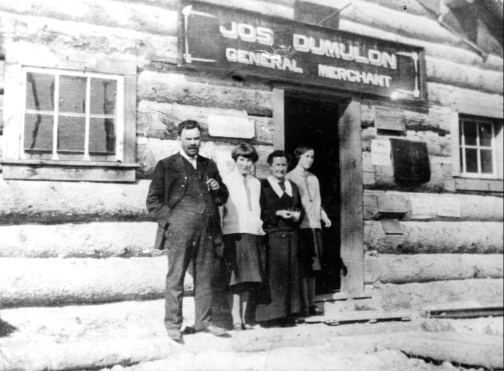 Black-and-white photograph of four people before a log front and a sign that reads, Jos Dumulon General Merchant.