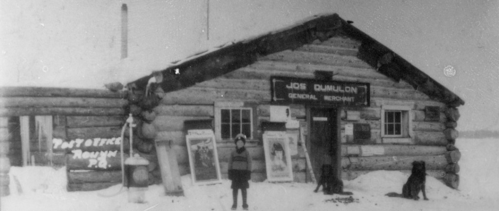 Black-and-white photograph of a log cabin with a sign that reads, Jos Dumulon General Merchant. In front, you can see a young boy with two dogs.