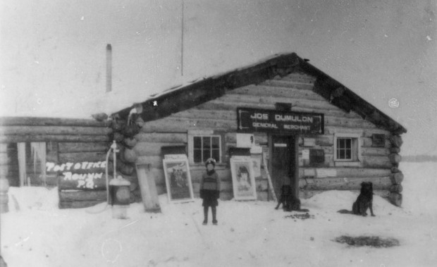 Black-and-white photograph of a log cabin with a sign that reads, Jos Dumulon General Merchant. In front, you can see a young boy with two dogs.