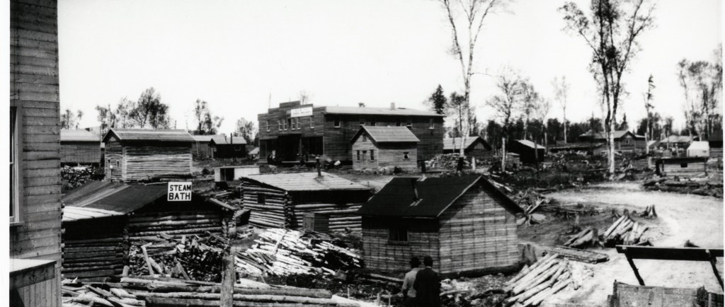 Black-and-white photograph of several rudimentary buildings made of wooden planks or timbers. One of them is a steam bath, identified as such on a sign.