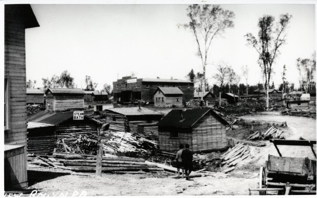 Black-and-white photograph of several rudimentary buildings made of wooden planks or timbers. One of them is a steam bath, identified as such on a sign.