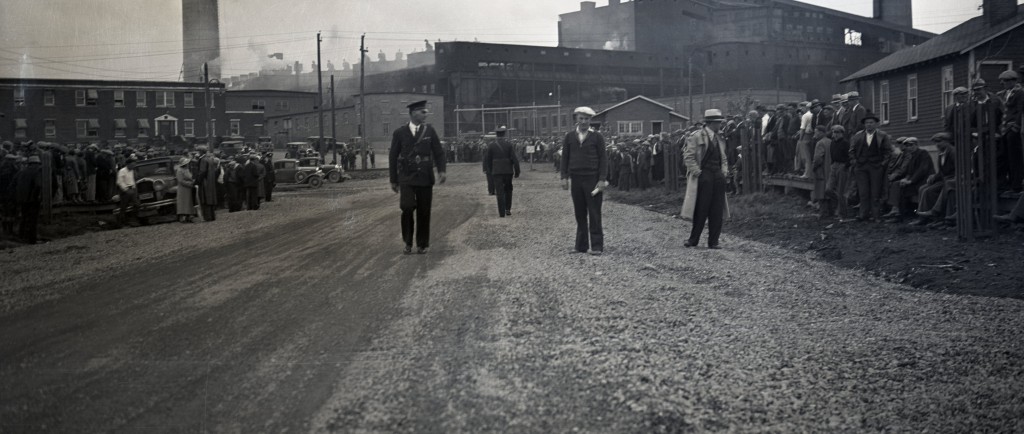 Black-and-white photograph of a road with people on opposite sides and five police officers in the middle, two of whom are wearing uniforms. You can see the smelter in the background.