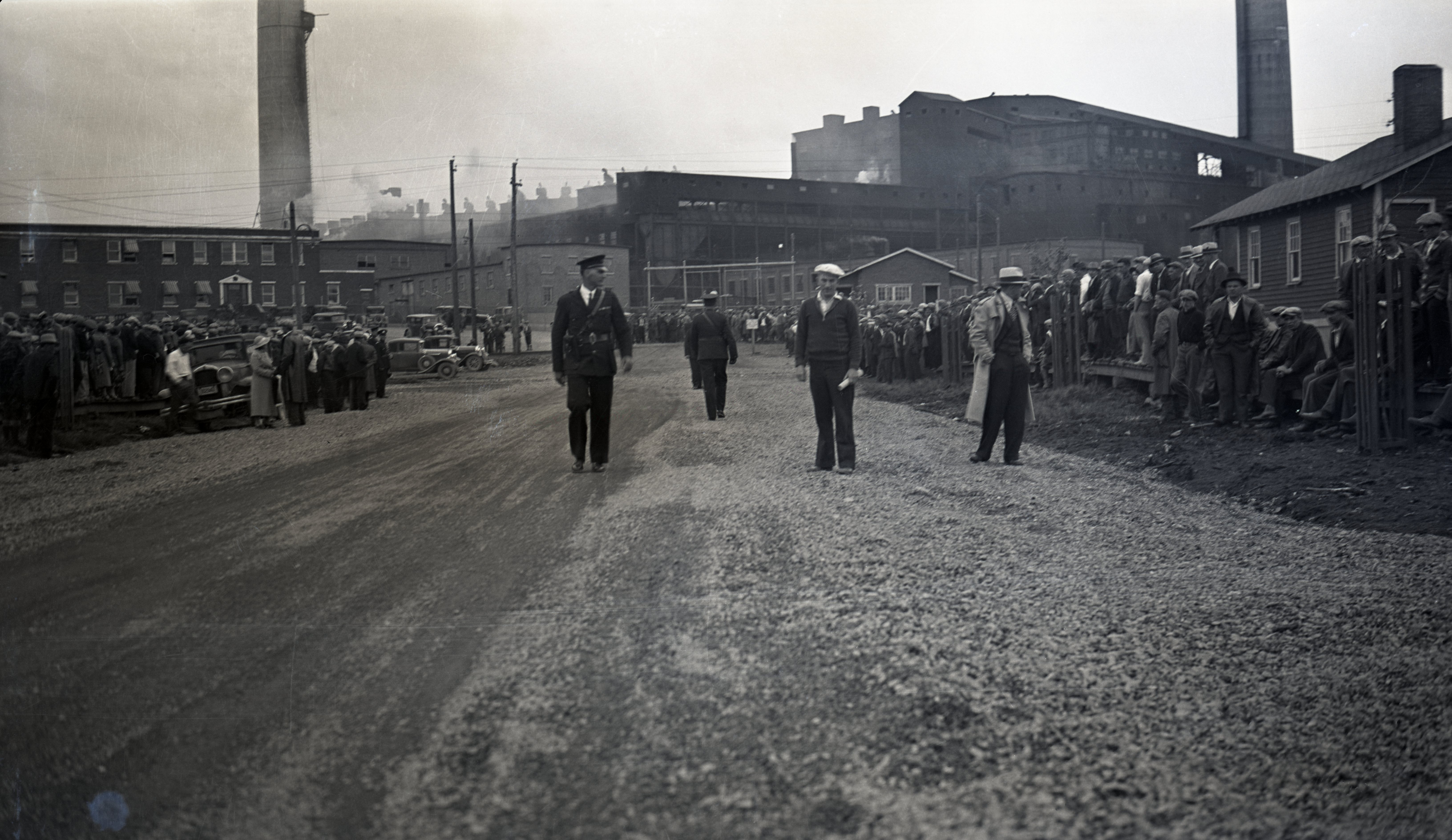 Black-and-white photograph of a road with people on opposite sides and five police officers in the middle, two of whom are wearing uniforms. You can see the smelter in the background.