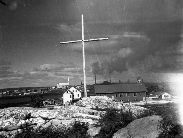 Black-and-white photograph of rocks, a cross, a church and a rectory. In the background, you can see Rouyn city on the left and the Horne mine facilities on the right.