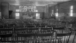 Photographie en noir et blanc de mauvaise qualité d’une église vide, dont la sacristie et la nef sont seulement séparées par un treillis de bois.