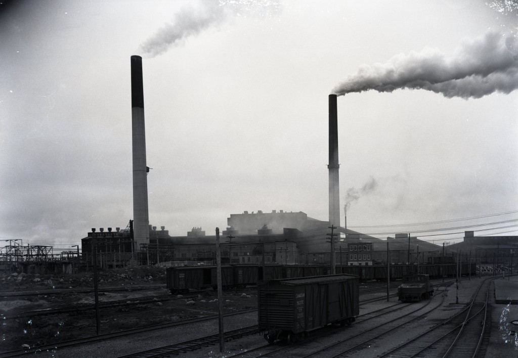 Photographie en noir et blanc d’un panorama de la fonderie. À l’avant-plan, plusieurs voies de chemin de fer et une dizaine de wagons identifiés au Canadian Pacific.