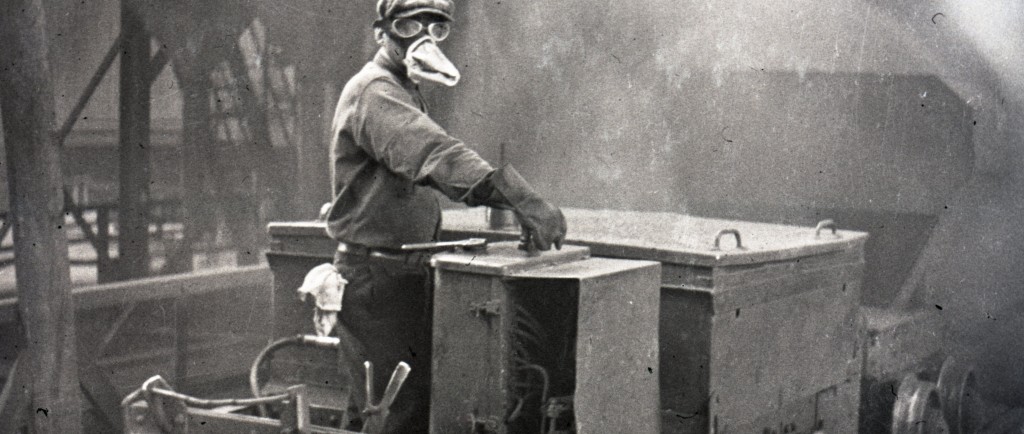 Photographie en noir et blanc d'un ouvrier portant un masque protecteur et des lunettes conduit un wagon à l’intérieur de la fonderie de la mine Horne. La présence de gaz rend la photo plutôt floue, mais on perçoit des structures en acier.