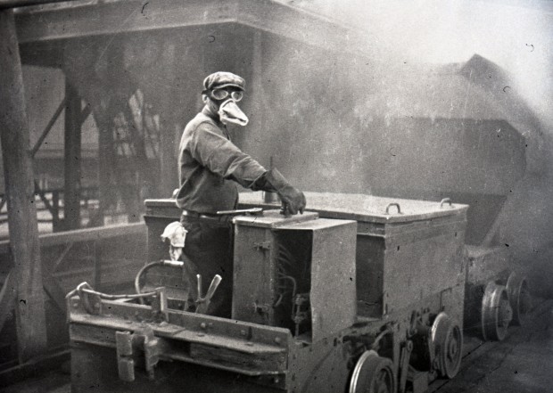 Black-and-white photograph of a worker wearing a protective mask and safety goggles while driving a car inside the Horne mine’s smelter. The picture looks blurred because of the gases, but you can see the steel structures.