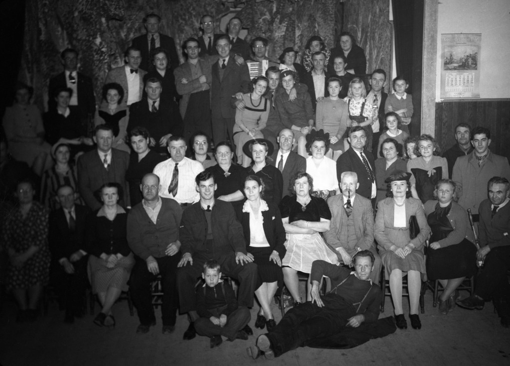 Photographie en noir et blanc d’une cinquantaine de personnes : vingt-trois femmes, vingt-quatre hommes et quelques enfants. Une des personnes tient un accordéon dans ses mains.