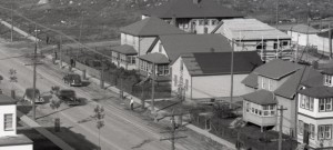 Black-and-white photograph of five adjacent buildings made of wooden planks. There are several cars travelling and people walking on the sidewalks.
