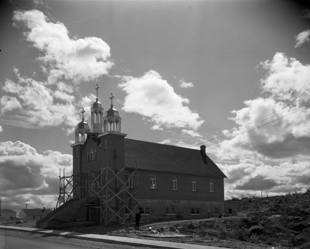 Black-and-white photograph of a church with three steeples, three crosses and a front enclosed within a scaffold. In the foreground, you can see steps leading to the main doors.
