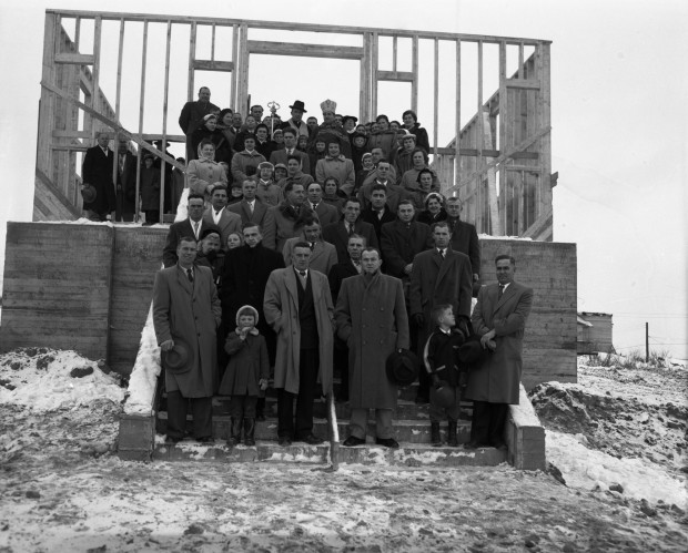 Black-and-white photograph of about 70 people on stairs in front of a wood structure. There are two Catholic priests, a Roman priest and a Ukrainian priest.