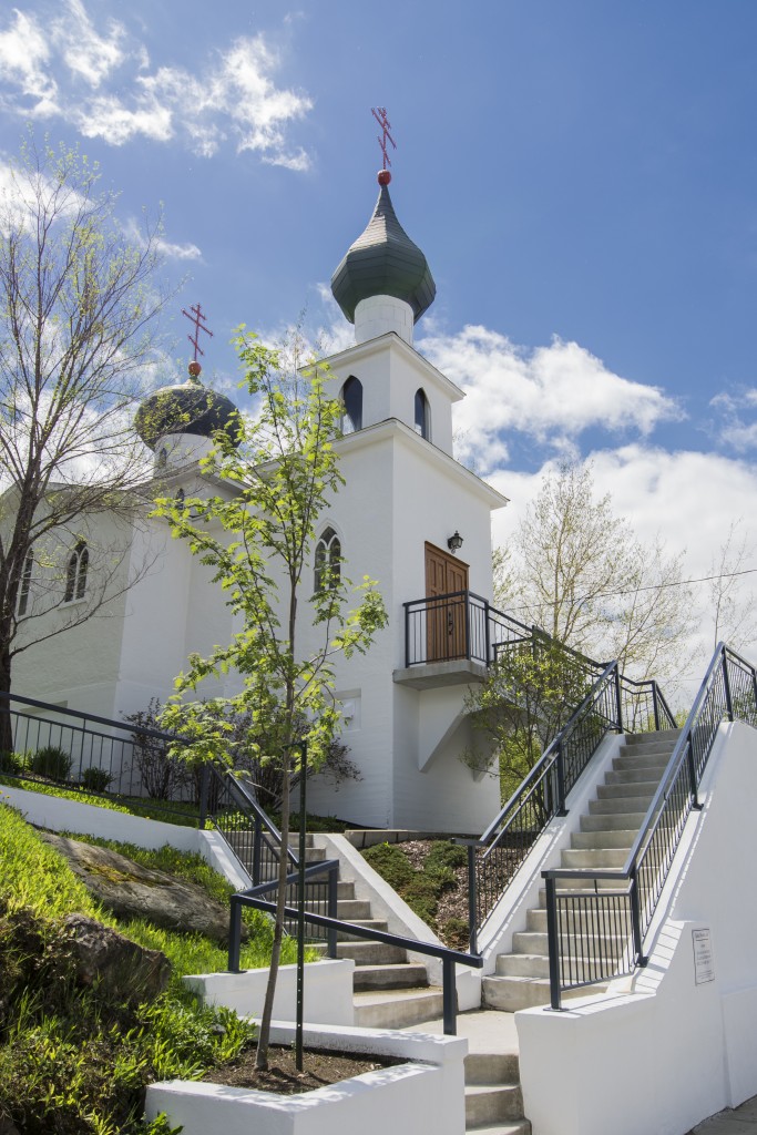 Colour photograph of a white church with orthodox crosses overlooking two bulb-shaped steeples. Concrete stairs lead to the main door and to the back of the church.