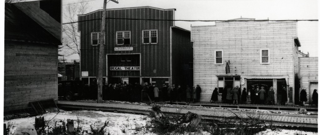 Photographie en noir et blanc de deux bâtiments avec des façades Boomtowns, dont un qui a une affiche Regal Theatre. Une foule attend sur le trottoir de bois devant l’édifice.