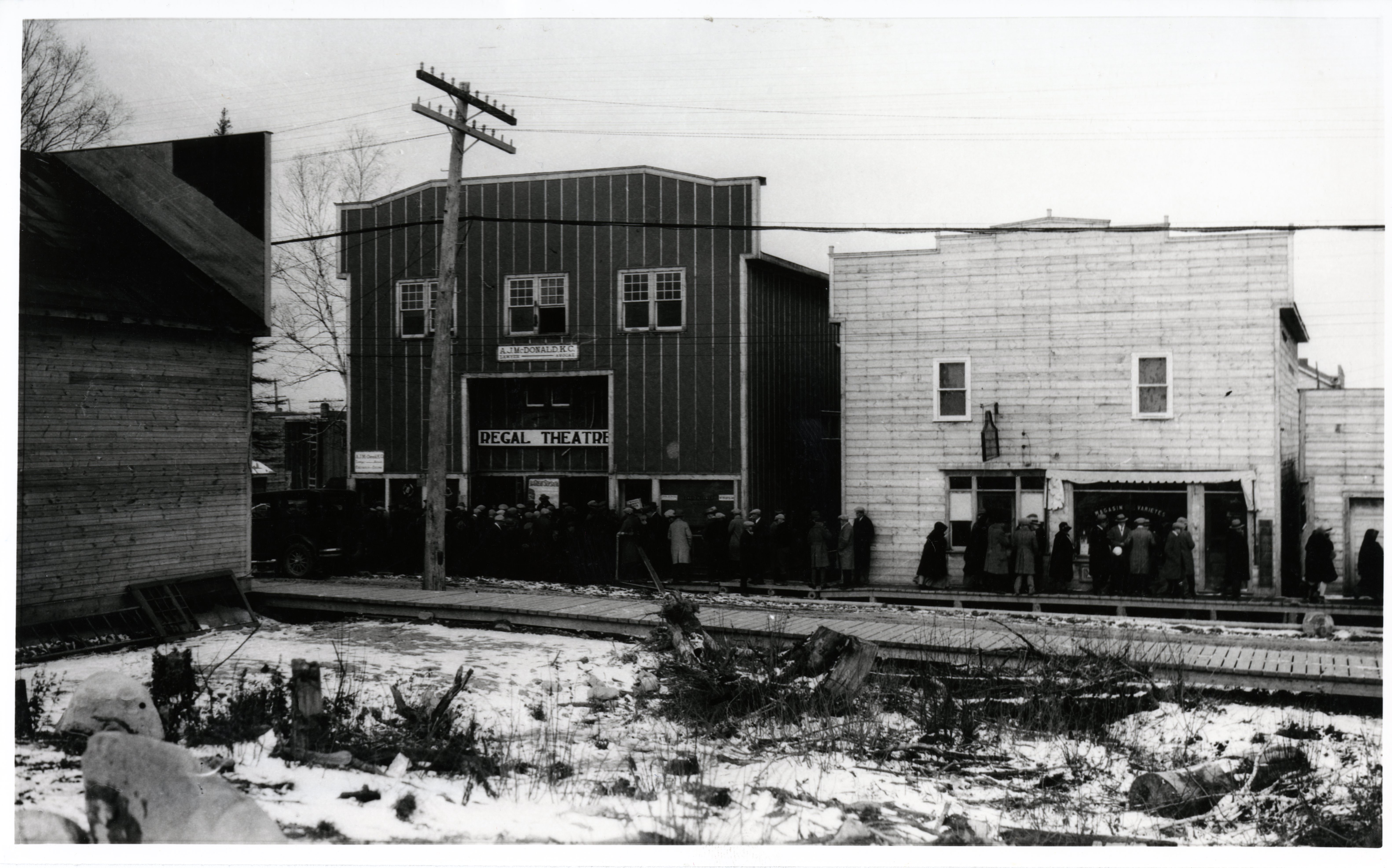 Black-and-white photograph of two buildings with boomtown-styled fronts. On one of them, there is a sign where it is written Regal Theatre. A crowd waits on the boardwalk in front of the building.