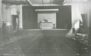 Black-and-white photograph of a theatre with both fixed and moveable chairs. On the stage there is an altar on which lies a tabernacle.