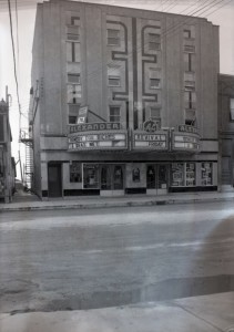 Black-and-white photograph of a four-story building with a theatre on the ground floor. You can see which movies were playing by reading the titles displayed at the entrance.