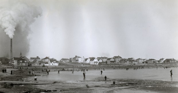 Black-and-white photograph of a snowless frozen lake on which there is about 40 ice skaters. You can see Noranda city and a smelter chimney in the background.