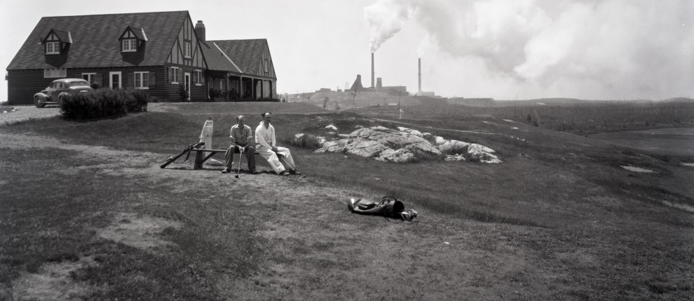Photographie en noir et blanc de deux hommes assis devant le pavillon du club de golf de Noranda avec, en arrière-plan, la mine Horne.