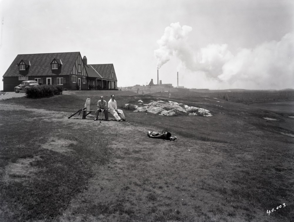 Black-and-white photograph of two men sitting in a lodge of the Golf Club of Noranda with the Horne mine in the background.