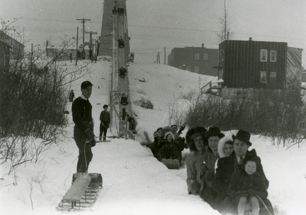 Black-and-white photograph of several people sliding on a wood structure installed on a hill.