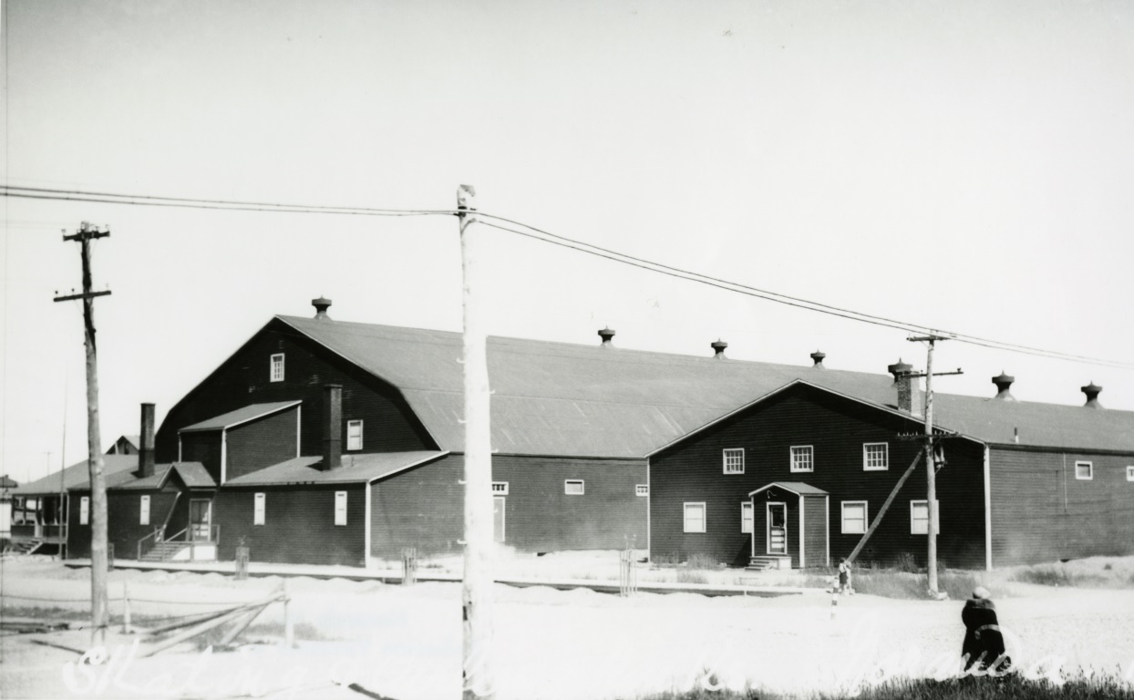 Black-and-white photograph of two buildings with tin roofs and very few windows. A blanket of snow covers the ground.