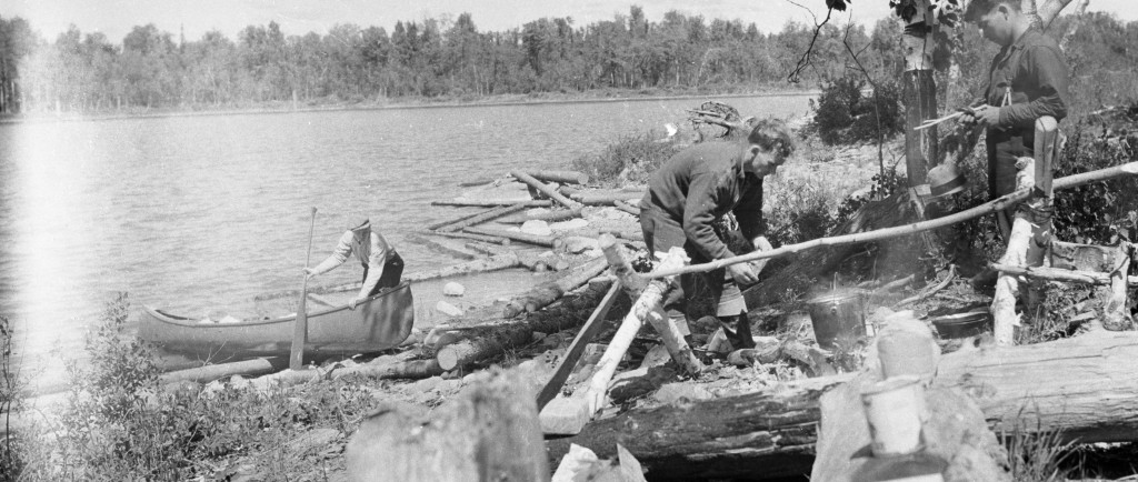 Black-and-white photograph of three prospectors. One of them is paddling a canoe while the two others are lighting a fire.