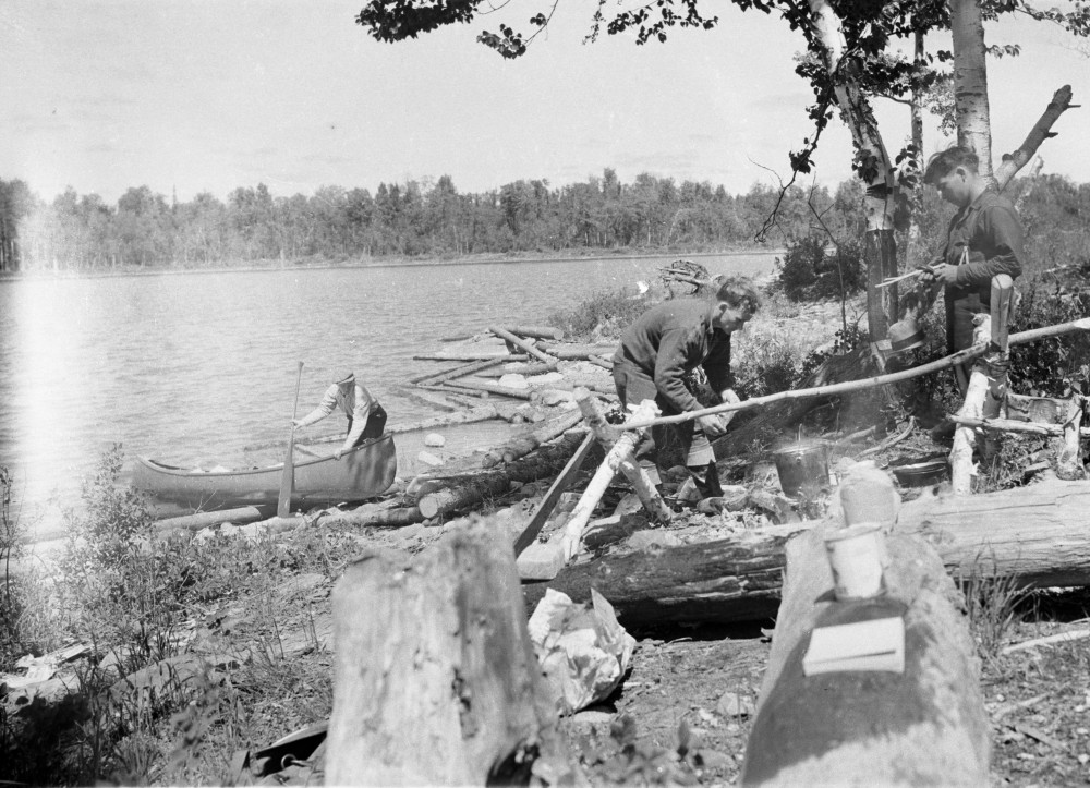 Black-and-white photograph of three prospectors. One of them is paddling a canoe while the two others are lighting a fire.