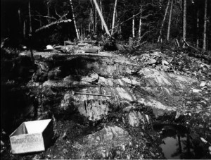 Black-and-white photograph of rock soils with two pickaxes and two wood box. There is a forest in the background.