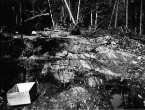 Photo en noir et blanc d’un sol rocheux, ainsi que de deux pioches et de deux boites de bois, avec la forêt en arrière-plan. 