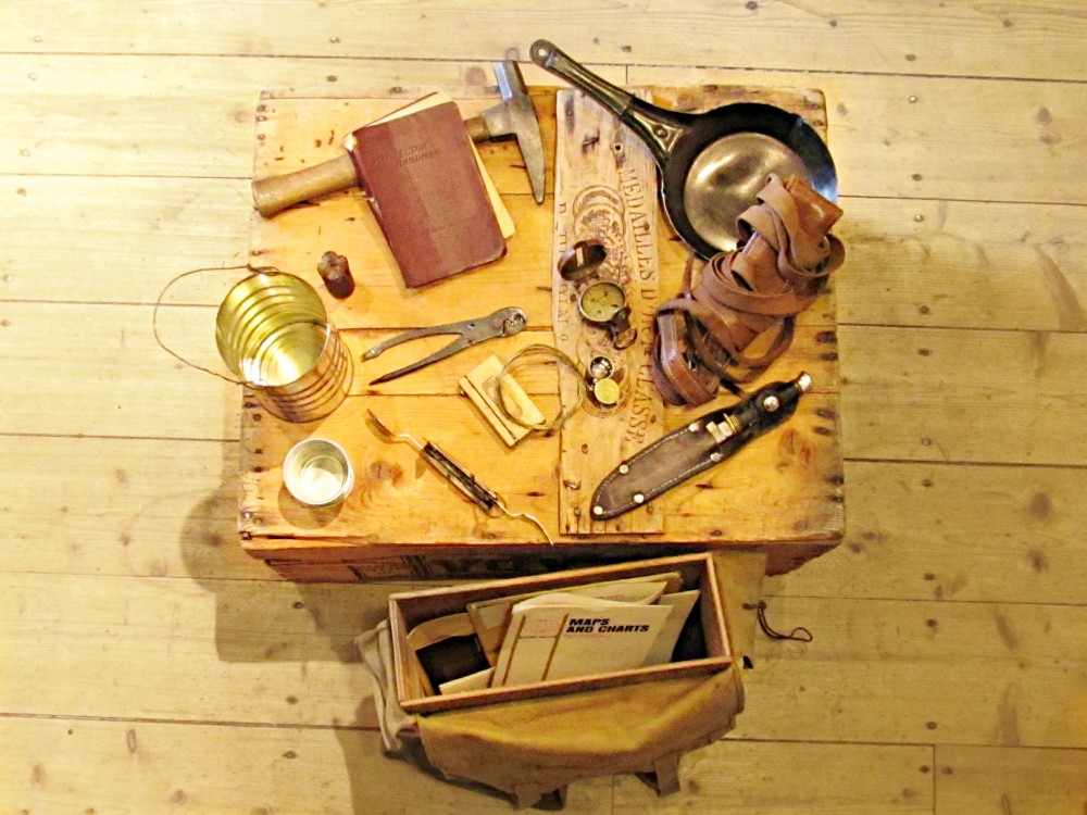 Colour photograph of about 15 prospecting artifacts spread out on a wood box and a canvas bag laying on the ground with a wood structure inside it. 