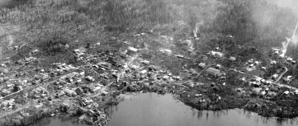 Photographie en noir et blanc d’une centaine de cabanes en bois rond installées de façon anarchique sur le bord du lac Osisko.